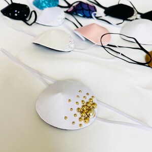 Könnte beinhalten: Eine Sammlung weißer Augenklappen mit verschiedenen Verzierungen, darunter Perlen, Pailletten und goldene Perlen. Die Augenklappen sind aus weichem Stoff gefertigt und haben ein elastisches Band für einen bequemen Sitz.