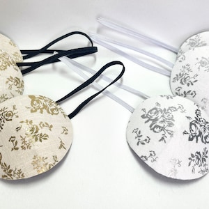 May include: Four eye patches with elastic straps. Two are cream-coloured with gold floral patterns, and two are white with silver floral patterns. The straps are black and white.