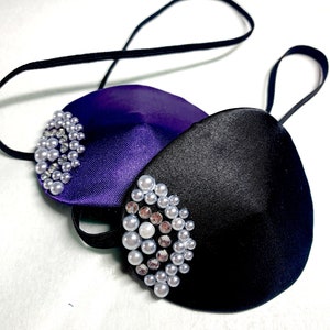 Puede incluir: Dos parches para los ojos con adornos de perlas y pedrería. Uno es morado y el otro es negro.