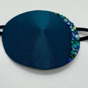 Könnte beinhalten: Türkisblaue Augenmaske aus Satin mit einer Verzierung aus blauen und grünen Perlen am Rand. Die Maske ist an einem schwarzen Gummiband befestigt.