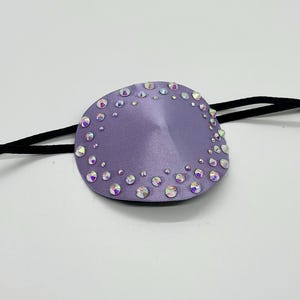 Puede incluir: Un parche para el ojo lavanda con pedrería iridiscente, sujeto a una banda elástica negra. El parche para el ojo tiene forma ovalada y tiene una textura similar a la del satén. La pedrería está dispuesta alrededor de los bordes y dispersa.