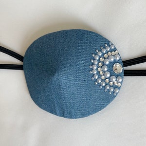 Puede incluir: Un parche para el ojo de mezclilla azul con un diseño decorativo de perlas blancas y diamantes de imitación.