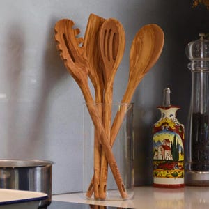 May include: A set of wooden kitchen utensils, including a slotted spoon, spatula, and solid spoon, presented in a clear glass. The utensils have a natural wood grain finish. A decorative oil bottle with a painted landscape design is visible in the background.