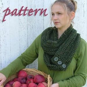 May include: A green knitted infinity scarf with three buttons on the side. The scarf is worn by a person holding a basket of red apples.