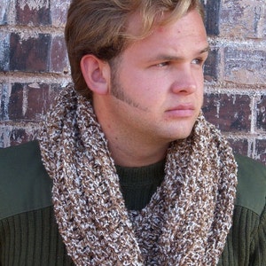 May include: A brown and white knit infinity scarf. The scarf is wrapped around the neck of a person wearing a green sweater. The scarf is made of a chunky knit material and has a textured appearance. The background is a brick wall.