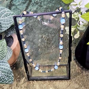 May include: A delicate necklace made with blue, gold, and black beads. The necklace is displayed in a glass box on a green surface.
