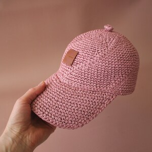 May include: A pink crocheted baseball cap with a brown leather patch. The cap has a curved brim and a slightly pointed crown.