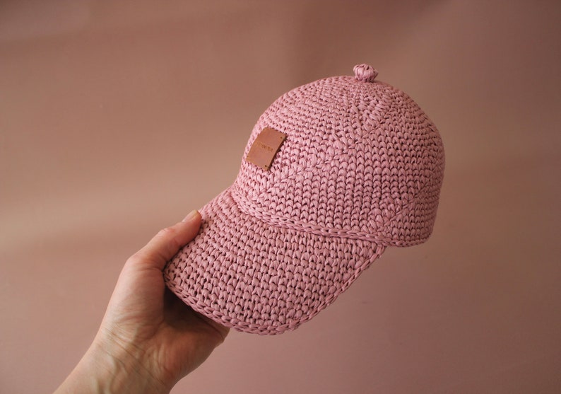 May include: A pink crocheted baseball cap with a brown leather patch. The cap has a curved brim and a slightly pointed crown.