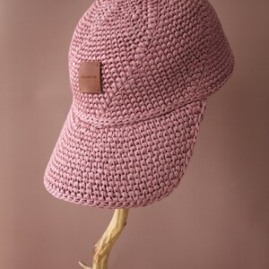 May include: A pink crocheted baseball cap with a brown leather label that says "ZHAMUNA". The cap is sitting on a wooden branch.