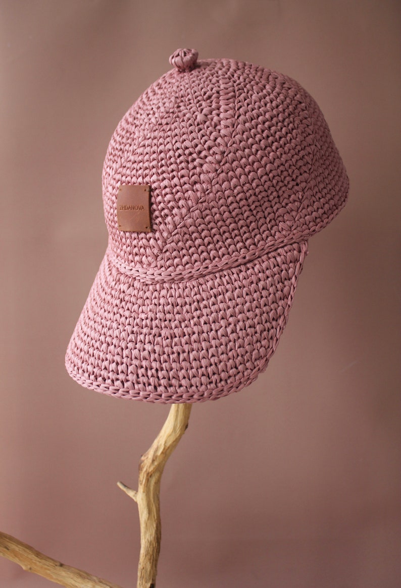 May include: A pink crocheted baseball cap with a brown leather label that says "ZHAMUNA". The cap is sitting on a wooden branch.