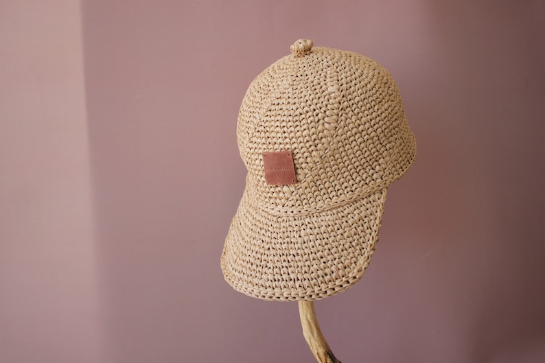 May include: A beige crocheted baseball cap with a brown leather patch on the front. The cap has a curved brim and is made of a natural material.