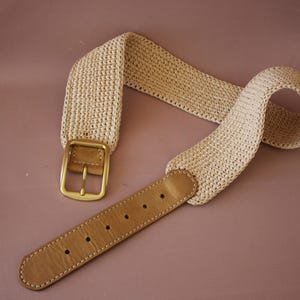 May include: A beige woven belt with a gold-colored buckle and a light brown leather strap. The belt is made of a textured, woven material and is designed to be worn around the waist. The belt is a neutral color and can be paired with various outfits.