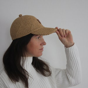 May include: A person wearing a light brown crocheted baseball cap with a brown leather patch on the front.