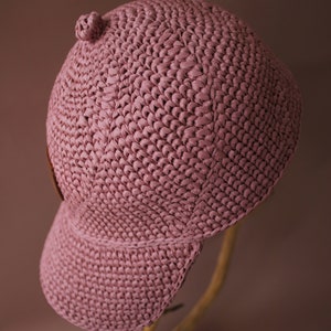 May include: A pink crocheted baseball cap with a curved brim. The cap is made of a soft, textured yarn and has a simple, classic design.