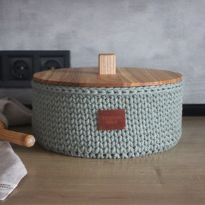 Op de afbeelding: Een groene gehaakte opbergcontainer met een houten deksel en een bruin leren label met de tekst "Handmade Home".