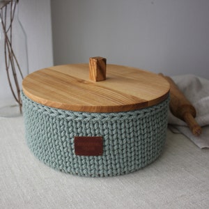 Może przedstawiać: Zielony koszyk na chleb wykonany na szydełku z drewnianym wiekiem i skórzaną etykietą. Koszyk stoi na lnianym obrusie.