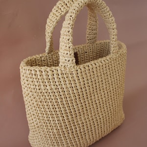 May include: A beige woven tote bag with two handles. The bag has a rectangular shape with a woven texture. The handles are also woven and attached to the top of the bag. The bag is made of a natural fiber material.