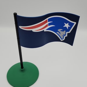 Könnte beinhalten: Eine Miniaturflagge mit dem Logo der New England Patriots. Die Flagge hat einen marineblauen Hintergrund mit dem Logo des Teams in Weiß, Rot und Blau. Sie ist auf einer schwarzen Stange mit einem grünen kreisförmigen Sockel montiert.