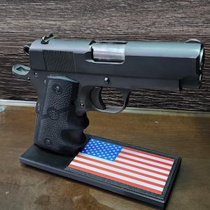 May include: A black handgun with a silver slide is displayed on a black stand with an American flag design. The stand is on a wooden surface.