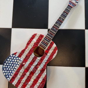 May include: Acoustic guitar decorated with red, white, and blue beads, creating an American flag design. The headstock features a star and chevron pattern. The guitar is set against a black and white checkered background.