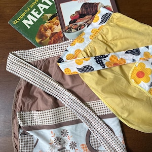 May include: A brown apron with a white and brown gingham waistband and a lower panel featuring a brown and white floral print, alongside a yellow apron with a white trim decorated with orange and black flower patterns and black polka dots.