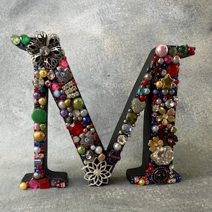 May include: Black wooden letter "M" decorated with an assortment of colorful beads, gems, and floral embellishments. The letter is adorned with various shapes and sizes of jewels, creating a textured and eye-catching design.