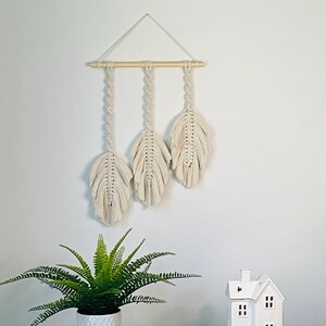 May include: A cream-coloured macrame wall hanging with three feather designs suspended from a natural wooden dowel. The feathers are intricately knotted, creating a textured, bohemian aesthetic. The piece is hung against a white wall, adding a touch of natural decor.