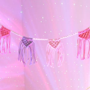 May include: A pink and purple macrame banner with triangle shaped flags and fringe. The banner is hanging against a pink sparkly background.