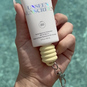 May include: White tube of Supergoop! Unseen Sunscreen SPF 50, 1.7 fl oz. The tube has a yellow, textured, spiral-shaped keychain attachment. The background is a blurred turquoise water.