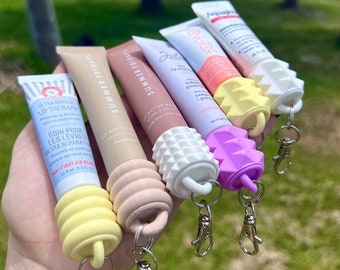 Puede incluir: Cinco tubos de bálsamo labial diferentes con llaveros adjuntos. Los tubos son de diferentes colores y tienen diferentes marcas. Los bálsamos labiales están en una mano, con un fondo borroso de una playa y palmeras.