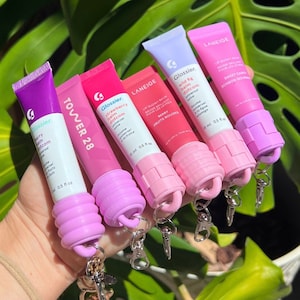 May include: Five different lip balm tubes with colourful labels and silicone keychain attachments. The tubes are labelled with the brand names Glossier, Tower 28, and Laneige. The lip balms are in shades of pink, purple, and white.