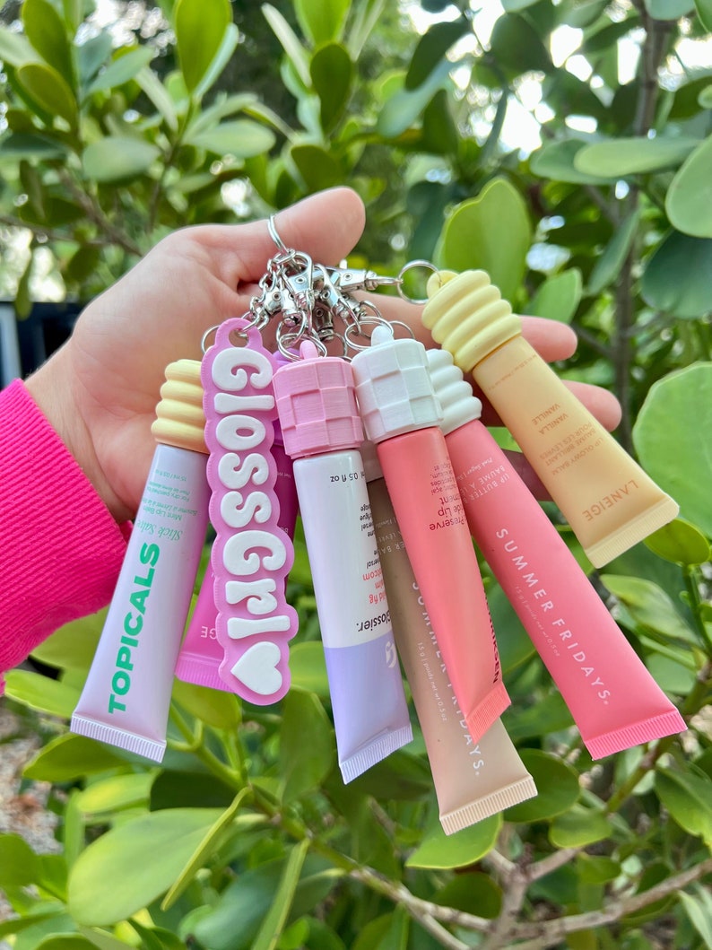 May include: A collection of five lip balm tubes with different brand names and colours. The tubes are attached to a keychain with a pink charm that says "Gloss Girl".