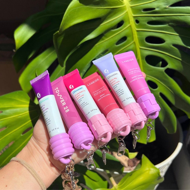 May include: Five different lip balm tubes with colourful labels and silicone keychain attachments. The tubes are labelled with the brand names Glossier, Tower 28, and Laneige. The lip balms are in shades of pink, purple, and white.