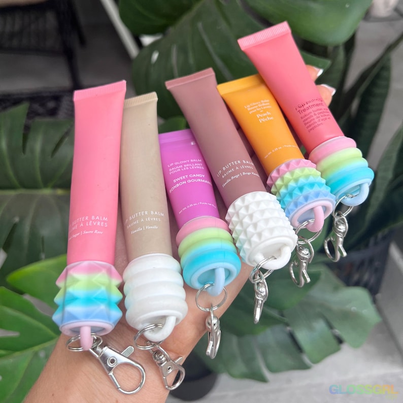 May include: Six colourful lip butter balms with different scents and flavours. Each balm has a white silicone cap with a colourful silicone ring and a keychain attached. The balms are arranged in a hand, with the pink balm at the bottom and the peach balm at the top.