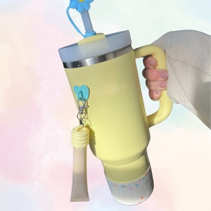 May include: A yellow stainless steel travel mug with a blue flower lid, a blue heart charm, and a clear tube with a white label. The mug has a confetti design on the bottom.
