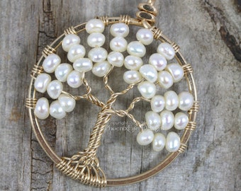 Puede incluir: Un colgante de árbol de la vida envuelto en alambre dorado con perlas blancas. El colgante está sobre una superficie de madera.