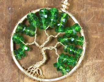 Puede incluir: Un colgante de árbol de la vida envuelto en alambre dorado con cuentas de vidrio verde. El colgante está sobre una superficie de madera.