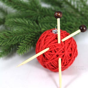 Puede incluir: Bola de adorno de lana roja con tres agujas de tejer de madera que sobresalen. El adorno est&aacute; sobre una superficie blanca con una rama de pino verde en el fondo. Copyright 2017 by K. Turner Phoenixfire Designs.