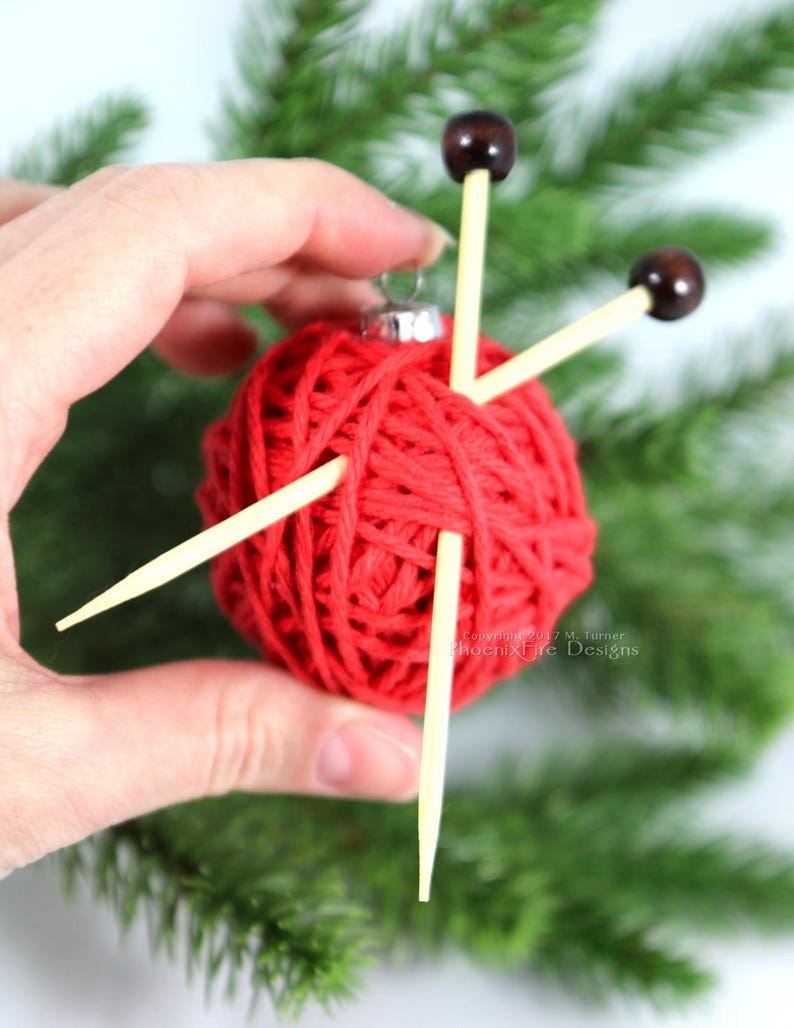 Puede incluir: Adorno navide&ntilde;o de bola roja de hilo con tres agujas de tejer de madera que sobresalen. El adorno se sostiene en una mano sobre un fondo verde.