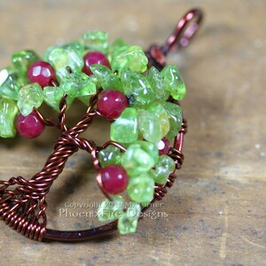 以下が含まれることがあります： 緑と赤の宝石ビーズが付いた銅線製の生命の木ペンダント。ペンダントは木製の表面にあります。Copyright 2013 M. Turner Phoenix Fire Designs.