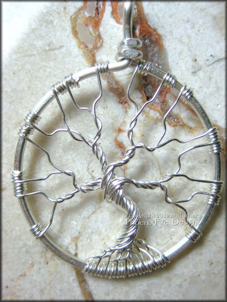 May include: A silver wire tree of life pendant with a circular design. The pendant is made of wire that is twisted and shaped to resemble a tree with branches and leaves. The pendant is on a white background.