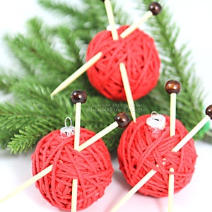 Puede incluir: Tres adornos de bola de hilo rojo con agujas de tejer de madera. Los adornos est&aacute;n decorados con cuentas de madera marr&oacute;n y cuelgan de una rama de pino verde.