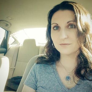 May include: A woman with long brown hair is wearing a grey t-shirt and a silver necklace with a blue tree pendant. She is sitting in the driver's seat of a car.