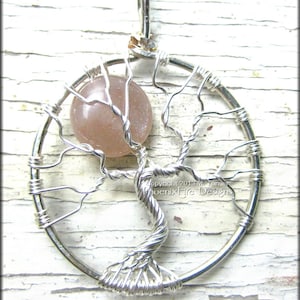 May include: Silver wire wrapped tree of life pendant with a pink gemstone. The pendant is on a white wooden background. Copyright 2013 M. Turner PhoenixFire Designs.