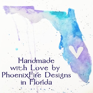 May include: A watercolour painting of the state of Florida with a white heart in the centre. The painting is in shades of blue and purple with white splatters. The text "Handmade with Love by PhoenixFire Designs in Florida" is written below the painting.