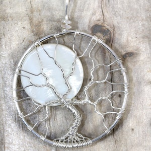 May include: A silver wire wrapped tree of life pendant with a white mother of pearl moonstone. The pendant is on a wooden background.