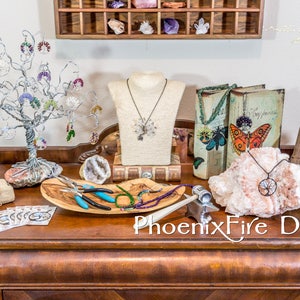May include: A selection of jewellery pieces are displayed on a wooden table, including necklaces, earrings, and bracelets. There are also tools for making jewellery, such as pliers and wire cutters. The table is decorated with a wooden tree, a book, and a crystal. The text "PhoenixFire Designs" is visible on the table.