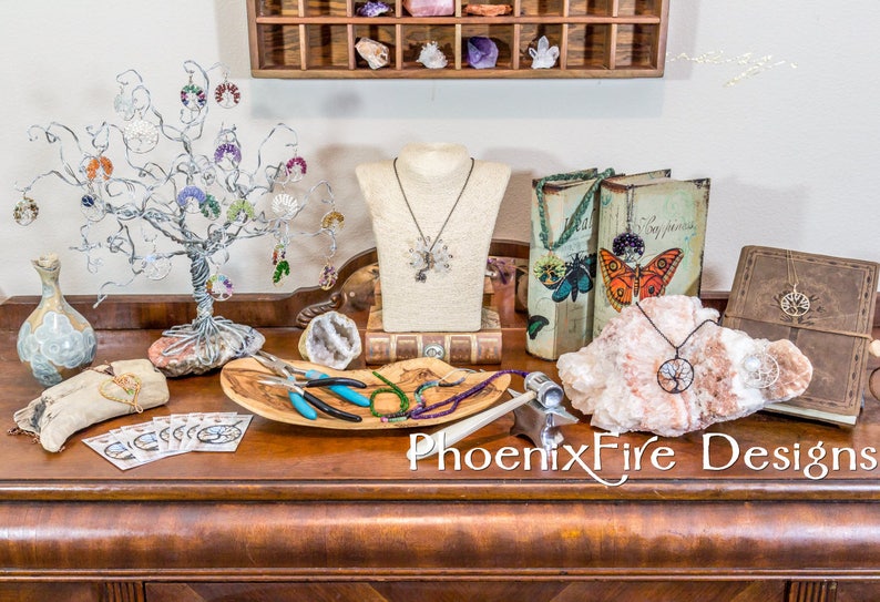May include: A selection of jewellery pieces are displayed on a wooden table, including necklaces, earrings, and bracelets. There are also tools for making jewellery, such as pliers and wire cutters. The table is decorated with a wooden tree, a book, and a crystal. The text "PhoenixFire Designs" is visible on the table.