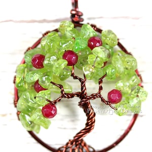 May include: A copper wire tree of life pendant with green and red gemstone leaves. The pendant is on a white wooden background. Copyright 2013 M. Turner Phoenix Fire Designs.