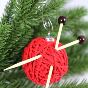 Puede incluir: Adorno navide&ntilde;o de bola de lana roja con dos agujas de tejer de madera que sobresalen. El adorno est&aacute; colgado de una rama de pino verde.